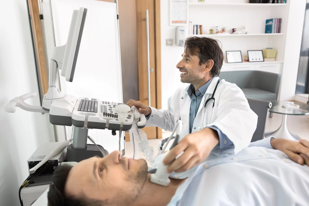 A grinning endocrinologist performs a thyroid ultrasound on a male patient.